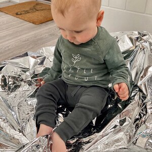 May include: A baby wearing a green long-sleeved top with the text "Little Scout" and black trousers sits on a silver blanket.
