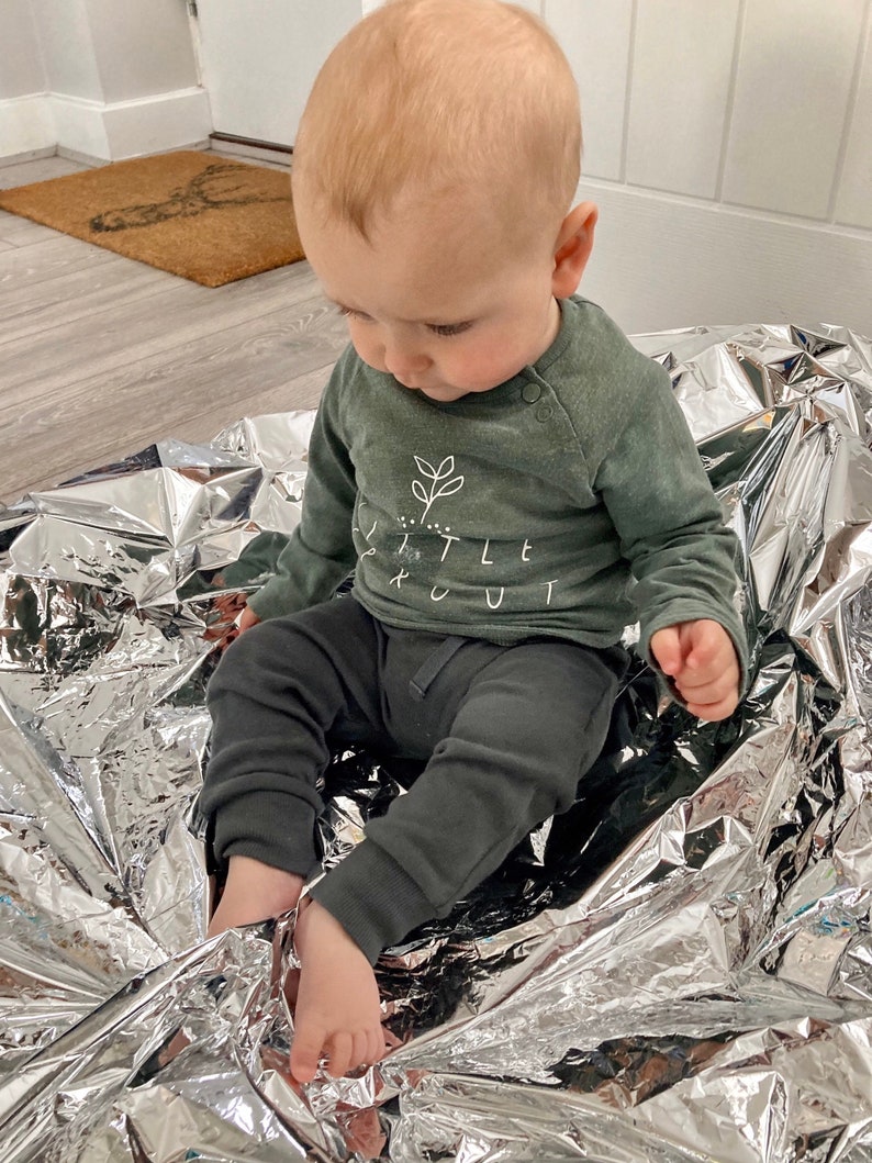 May include: A baby wearing a green long-sleeved top with the text "Little Scout" and black trousers sits on a silver blanket.