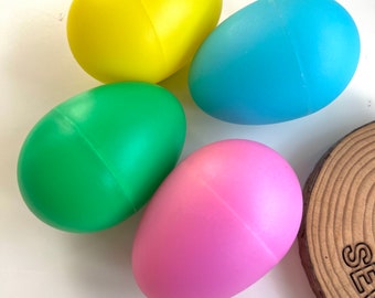 May include: Four plastic eggs in different colors: yellow, blue, green, and pink. The eggs are arranged in a circle on a white surface.