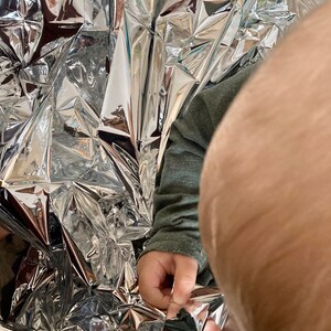 May include: A child's hand reaches into a silver emergency blanket, pulling it apart. The blanket is crumpled and reflective.