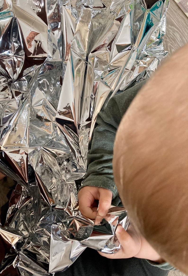 May include: A child's hand reaches into a silver emergency blanket, pulling it apart. The blanket is crumpled and reflective.