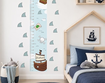 May include: A vertical wall decal height chart featuring a light blue ocean background with scattered shark fins, a treasure chest, a map, a skull and crossbones, and a brown pirate ship with the name Jackson printed on the sail.