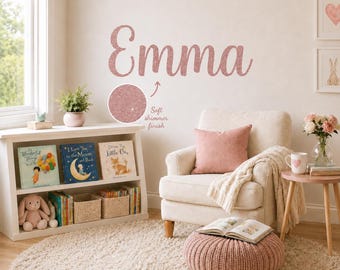 May include: A wall decal featuring the name Emma in a cursive script font with a rose gold glitter finish. A circular inset shows a close-up of the textured, shimmering surface, accompanied by the text Soft shimmer finish with an arrow.
