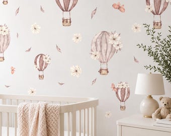 May include: A set of wall decals featuring watercolor-style hot air balloons in shades of brown and beige, decorated with white flowers and butterflies, scattered across a wall above a white crib and a white bedside table with a lamp.