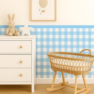 Op de afbeelding: Een kinderkamer met een witte commode, een wieg van riet en een ingelijste afbeelding van een luchtballon. De muur heeft een blauw-wit ruitpatroon. Een pluchen konijn en een sterdecoratie staan op de commode.