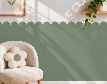 May include: A sage green scalloped wall decal border applied to a white wall, featuring a rounded edge design. A cream-colored plush flower-shaped pillow sits on a matching cream-colored armchair, with a framed flower print and a hanging plant above.