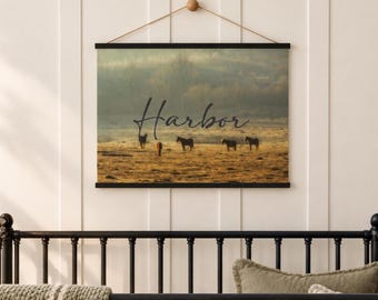 May include: A rectangular wall hanging featuring a landscape photograph of four horses in a golden field under a misty sky. The word Harbor is printed in black cursive script across the center. The print is held by black wooden rails and a string.