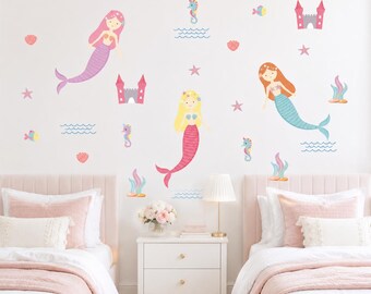 May include: A set of wall decals featuring three mermaids with pink, red, and blue tails, two pink castles, seahorses, starfish, seashells, and seaweed. The illustrations are colorful and arranged on a wall above two beds with pink bedding.