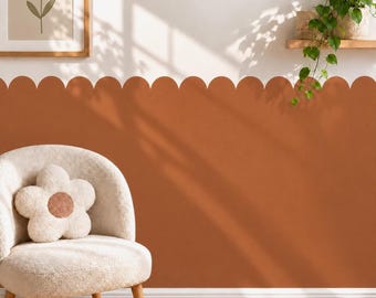May include: A scalloped wall decal in a solid terracotta color applied to the lower half of a wall. The wall features a white chair with a flower-shaped pillow, a framed flower print, and a floating shelf with a green trailing plant.