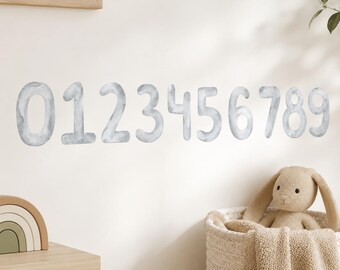 May include: A set of wall decals featuring the numbers zero through nine in a light gray watercolor texture. The numbers are arranged in a horizontal line on a plain wall, with a beige plush rabbit toy in a woven basket visible below.