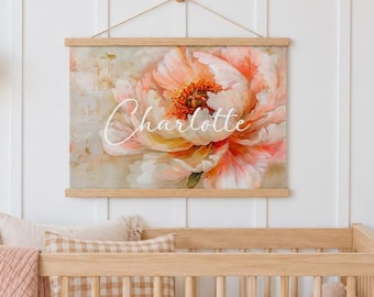 May include: A rectangular wall hanging featuring a large, soft-focus peach and white peony flower illustration. The name Charlotte is printed in white cursive script across the center of the bloom. The artwork is held by light wood rails.