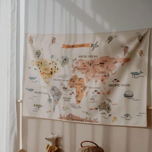 May include: A rectangular wall tapestry featuring a world map with a cream-colored background. The continents are illustrated in muted earth tones and labeled with ocean names. The map includes small drawings of animals, ships, and hot air balloons.