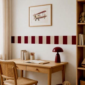 Könnte beinhalten: Ein Schreibtisch aus Holz mit einem Stuhl mit Rohrgeflecht, einer roten pilzförmigen Lampe und einem Stiftehalter. Ein gerahmter Druck eines roten Doppeldeckers hängt über einer Wandbordüre mit dunkelroten und beigen vertikalen Streifen.