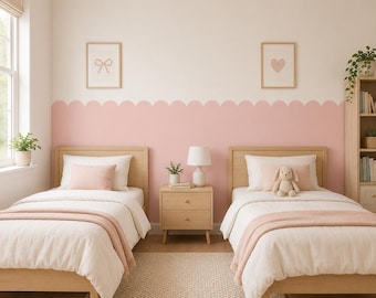May include: Two light wood twin beds with white bedding and pink throw blankets, separated by a light wood nightstand with a white lamp. The wall features a scalloped pink painted border, two framed prints of a bow and heart, and a tall bookshelf.