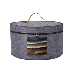May include: A gray felt hat storage case with a brown leather handle and a zippered closure. The case has a clear window showing several hats inside. The case is round and designed to protect hats from dust and damage.