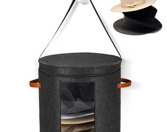 May include: Gray felt hat storage container with a clear window, hanging from a hook on a white wall. The container has brown leather handles and a zipper closure. The window shows a stack of hats inside.