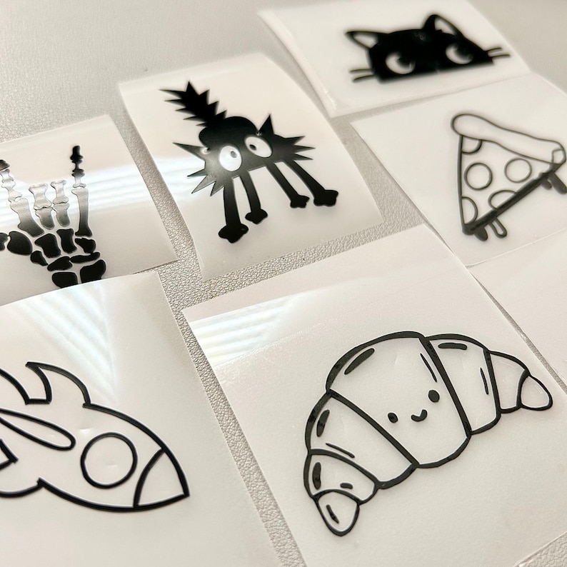 May include: A collection of black and white vinyl stickers featuring various designs. The stickers include a cat, a slice of pizza, a croissant, a rocket ship, a skeleton hand, and a spiky creature. The stickers are on a white background.