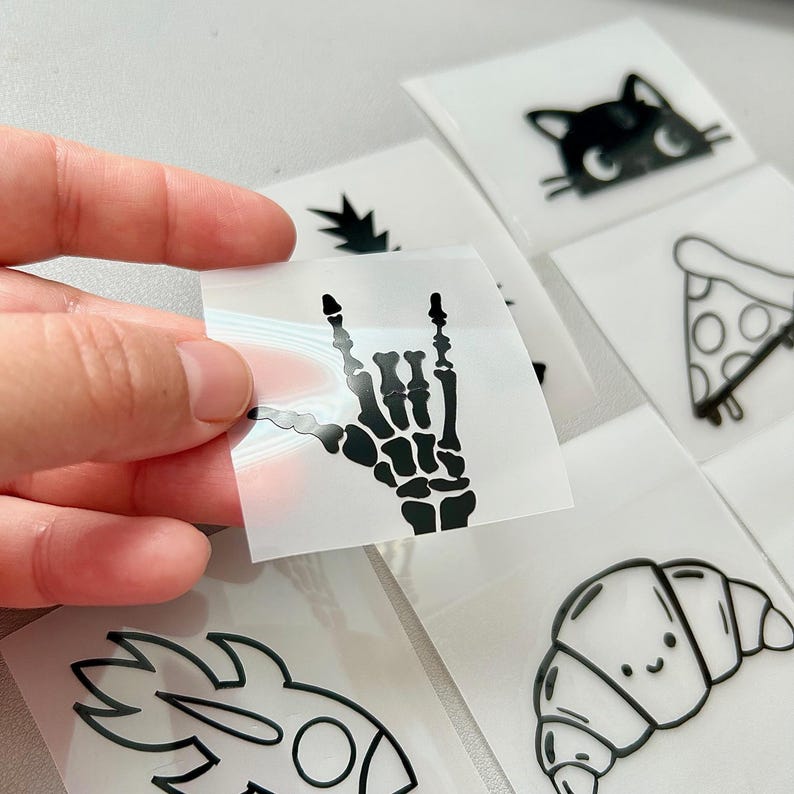 May include: A collection of black vinyl stickers featuring various designs. One sticker shows a skeleton hand making a rock and roll gesture. Other stickers include a cat face, a slice of pizza, a croissant, a pineapple, and a rocket.