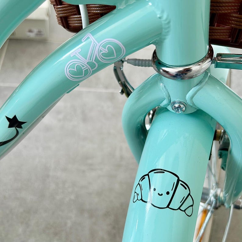 May include: Close-up of a light teal bicycle frame with a brown wicker basket. The frame has white decals of a bicycle and a star. A black and white cartoon croissant sticker is on the front fender.