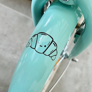 May include: A light blue bicycle fender with a black and white cartoon croissant sticker. The sticker has a smiling face. The bicycle has a silver metal fork and handlebars.
