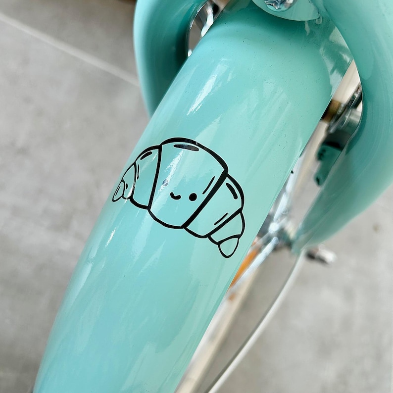 May include: A light blue bicycle fender with a black and white cartoon croissant sticker. The sticker has a smiling face. The bicycle has a silver metal fork and handlebars.