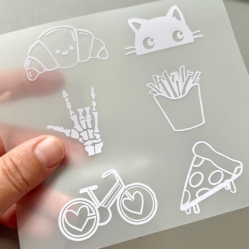 May include: White die-cut stickers featuring various designs, including a croissant, cat face, french fries, pizza slice, bicycle with heart-shaped wheels, and a skeleton hand making a rock-on gesture. The stickers are on a translucent background.