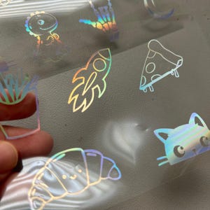 May include: A collection of holographic stickers featuring various designs. The stickers include a dinosaur, a rocket ship, a slice of pizza, a cat, a croissant, and a hand making a rock and roll gesture.