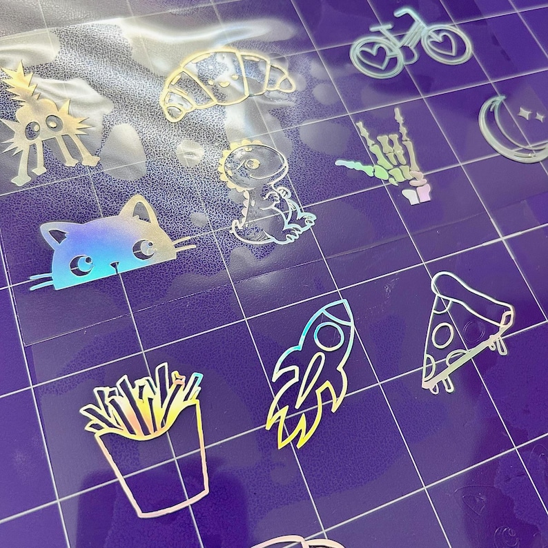 May include: A collection of iridescent stickers featuring various designs, including a cat, dinosaur, rocket ship, pizza slice, french fries, croissant, and a bicycle with a heart-shaped wheel. The stickers are on a purple grid background.
