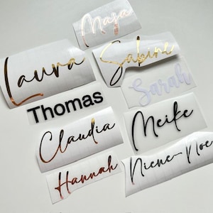 Könnte beinhalten: Eine Sammlung individueller Namensaufkleber in verschiedenen Farben und Schriftarten. Namen sind Laura, Maja, Sabine, Sarah, Thomas, Claudia, Meife, Hannah und Niene-Noe. Die Aufkleber sind auf weißem Hintergrund mit goldfarbenen, schwarzen und roségoldenen Buchstaben.