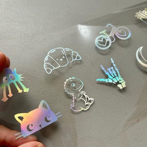 May include: Holographic stickers featuring various designs, including a cat, dinosaur, croissant, bicycle with heart-shaped wheels, and a skeleton hand. The stickers have a rainbow sheen and are on a clear backing.
