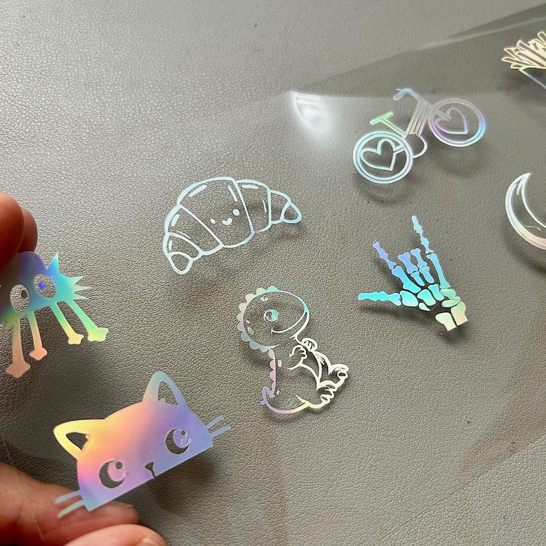May include: Holographic stickers featuring various designs, including a cat, dinosaur, croissant, bicycle with heart-shaped wheels, and a skeleton hand. The stickers have a rainbow sheen and are on a clear backing.