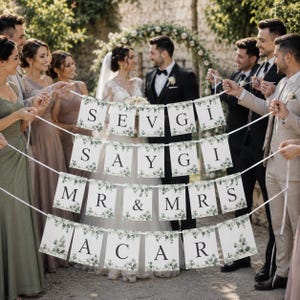 Könnte beinhalten: Ein Hochzeitsbanner mit weißen rechteckigen Flaggen, verziert mit grünen Eukalyptusblatt-Illustrationen und schwarzer Schrift, die SEVGI SAYGI MR & MRS ACAR lautet, gehalten von einer Hochzeitsgesellschaft in festlicher Kleidung.