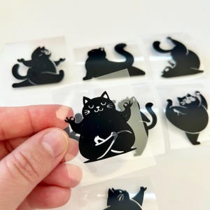 Könnte beinhalten: Schwarze Katzenaufkleber in verschiedenen Yoga-Posen. Die Aufkleber zeigen eine lächelnde Katze in verschiedenen Positionen, darunter der Lotussitz und Dehnübungen. Die Aufkleber befinden sich auf einer transparenten Rückseite und eignen sich ideal zum Basteln oder Dekorieren.