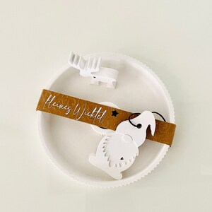 May include: White ceramic Christmas ornament featuring a reindeer, a gnome, and a brown tag that reads "Kleines Wichtel".