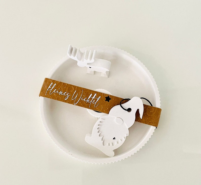 May include: White ceramic Christmas ornament featuring a reindeer, a gnome, and a brown tag that reads "Kleines Wichtel".