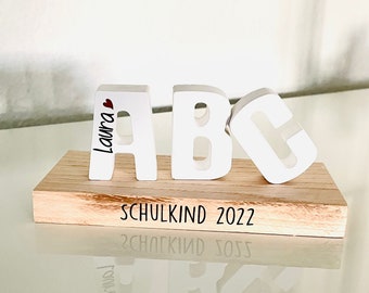 May include: White wooden letters spelling "ABC" with a heart and the name "Laura" on the "A". The letters are sitting on a wooden base with the text "SCHULKIND 2022" engraved on it.