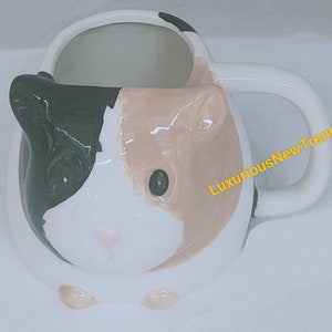 May include: White ceramic mug shaped like a guinea pig with black, brown, and pink painted details. The mug has a handle on the right side and the words "LuxuriousNewTrend" are printed on the handle.