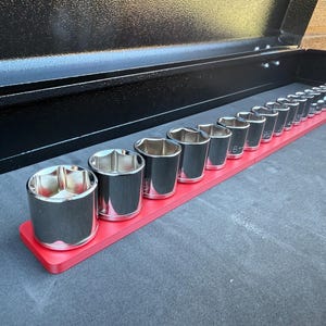 Könnte beinhalten: Ein Satz verchromter Steckschlüssel auf einem roten Halter in einer schwarzen Werkzeugkiste. Die Steckschlüssel sind in verschiedenen Größen erhältlich und für die Verwendung mit einem Schraubenschlüssel bestimmt. Die Werkzeugkiste ist geöffnet.