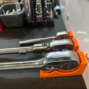 May include: Three silver ratcheting spanners are stored in orange plastic holders mounted on a white wall. The holders are labelled with the sizes 1/2, 3/8, and 1/4.