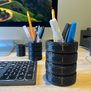Może przedstawiać: Czarne organizery biurowe w kształcie ułożonych w stos opon, trzymające długopisy, ołówki i markery. Organizery znajdują się na jasnym biurku obok klawiatury i monitora.