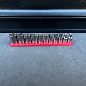 Könnte beinhalten: Ein Satz verchromter Steckschlüssel in verschiedenen Größen, angeordnet auf einem leuchtend roten Halter. Die Steckschlüssel sind zylindrisch mit sechseckiger Öffnung. Das Set wird vor einem dunklen Hintergrund präsentiert, was auf ein Werkzeug- oder Automobilthema hindeutet.