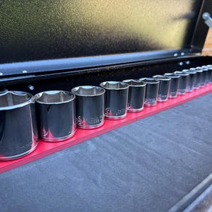 Könnte beinhalten: Ein Satz verchromter Steckschlüssel auf einem roten Halter in einer schwarzen Werkzeugkiste. Die Steckschlüssel haben verschiedene Größen, der größte befindet sich auf der linken Seite. Die Werkzeugkiste ist geöffnet und zeigt die geordneten Werkzeuge.