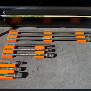 May include: A set of impact driver bits and sockets in various sizes. The tools are black with orange holders and blue accents. The text "LIBRATON IMPACT CR-V" is printed on the tools. The tools are arranged on a dark gray surface.