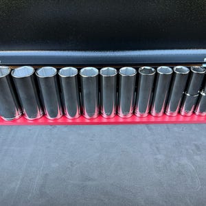 Könnte beinhalten: Ein Satz verchromter Steckschlüssel in verschiedenen Größen, angeordnet auf einem roten Halter. Die Steckschlüssel haben eine sechseckige Öffnung. Das Set wird vor einem dunklen Hintergrund präsentiert, was auf ein Werkzeugorganisationssystem hindeutet.