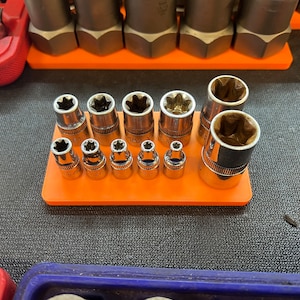 May include: A collection of silver metal sockets of various sizes, arranged on an orange tray. The sockets have star-shaped openings and are designed for use with a wrench. Other tools are visible in the background.