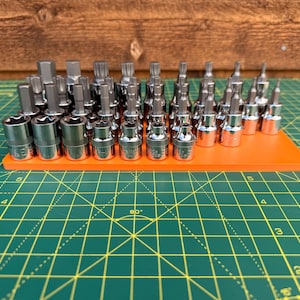 May include: A collection of silver-coloured metal socket bits and hex keys arranged in rows on a rectangular orange plastic organiser tray. The set includes various sizes of star-shaped, hexagonal, and spline-drive bits with metallic finishes.