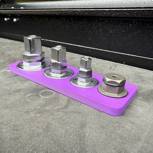 May include: A purple tool organizer holds four metal socket adapters. The adapters are various sizes and shapes, with a silver finish. The organizer is rectangular and sits on a gray surface, with a black object in the background.