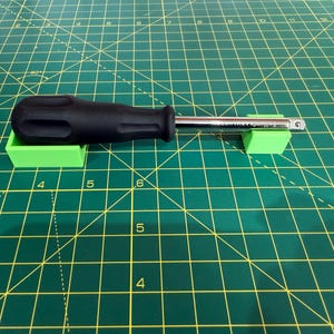 May include: A black-handled screwdriver with a silver shaft rests on two small, bright green rectangular supports. The tools are positioned on a green cutting mat with yellow grid lines and numbers.