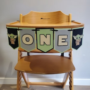 May include: Wooden high chair with a banner that reads "ONE" in white letters. The banner has green, gray, and black pennants with images of a character. The high chair is made of light-colored wood.