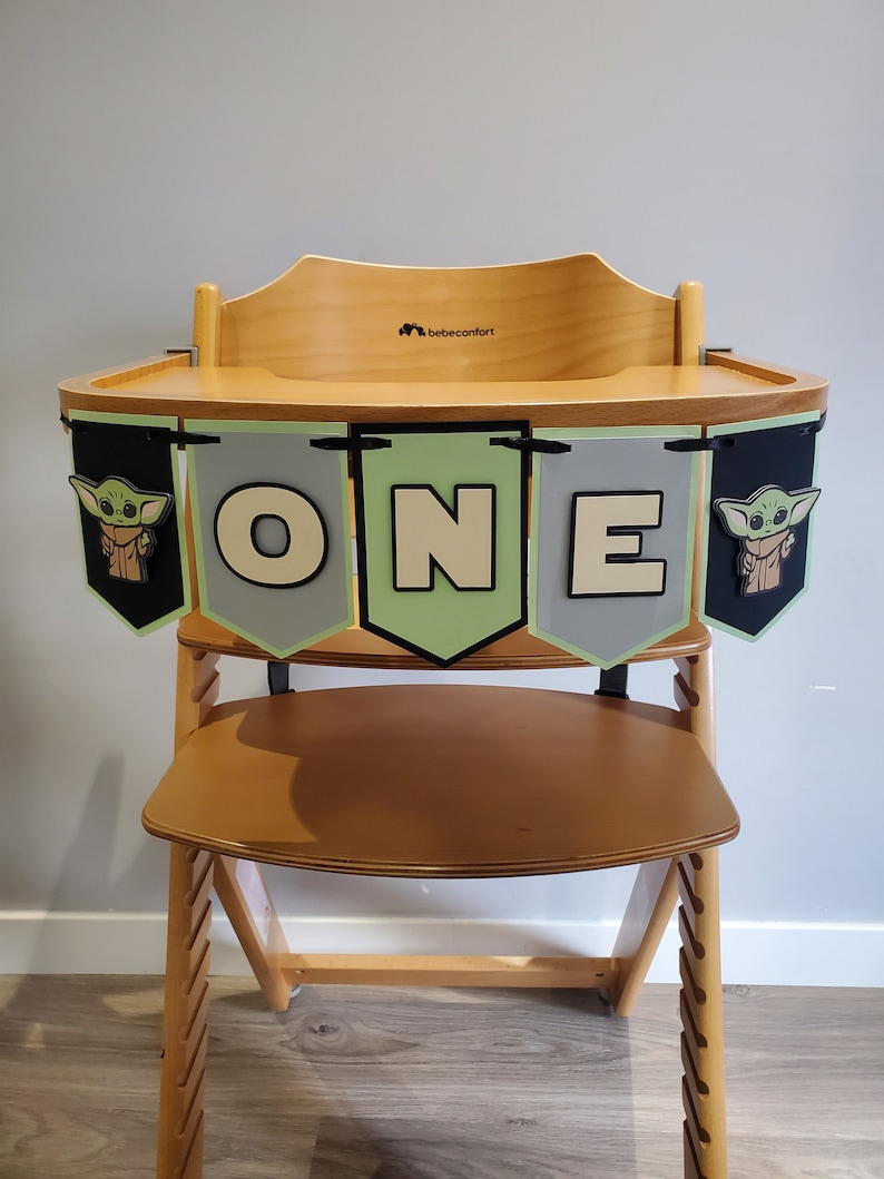 May include: Wooden high chair with a banner that reads "ONE" in white letters. The banner has green, gray, and black pennants with images of a character. The high chair is made of light-colored wood.